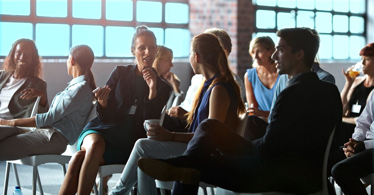 Two women talking in a room with more people while being sat waiting for a presentation to begin. 